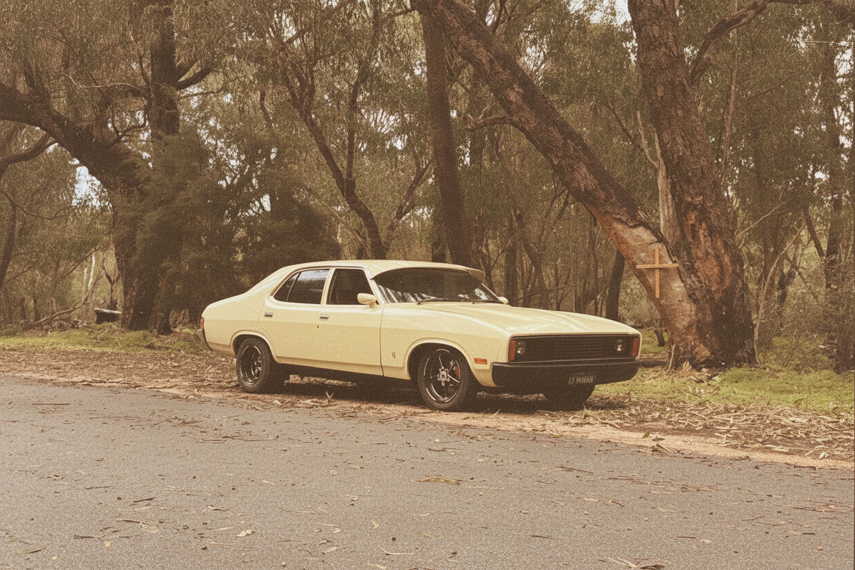 A pale yellow classic muscle car parked on the side of a paved road in a wooded outdoor setting with large trees and greenery.